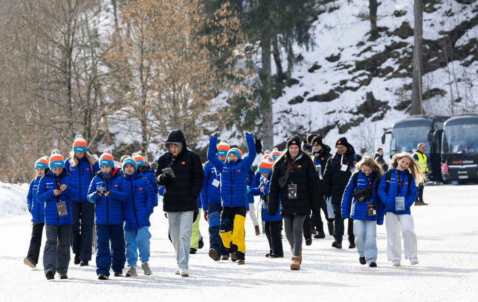 Kinder laufen als Gruppe zur Skiflug-Schanze. Dabei sieht man die Jacken und Mützen mit dem Mastercard Logo.